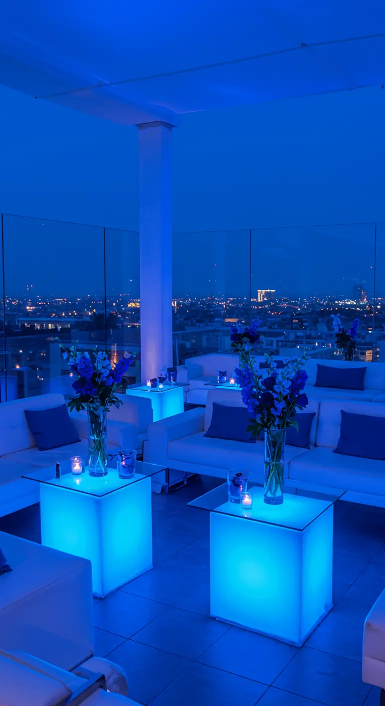 Fête sur toit-terrasse monochrome bleue avec cubes lumineux, canapés blancs et vue sur la ville.