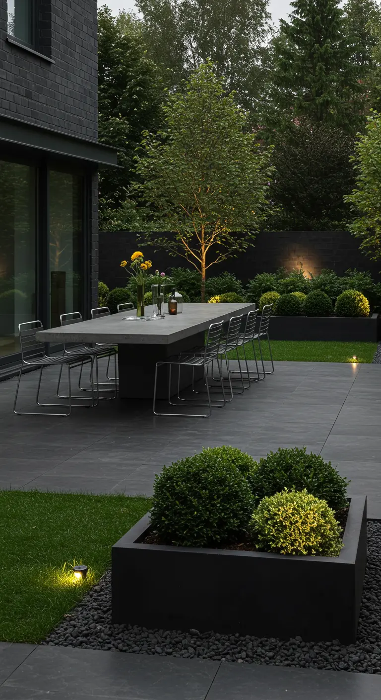 Longue table en béton et chaises en acier sur un patio sombre avec éclairage au sol et buis.