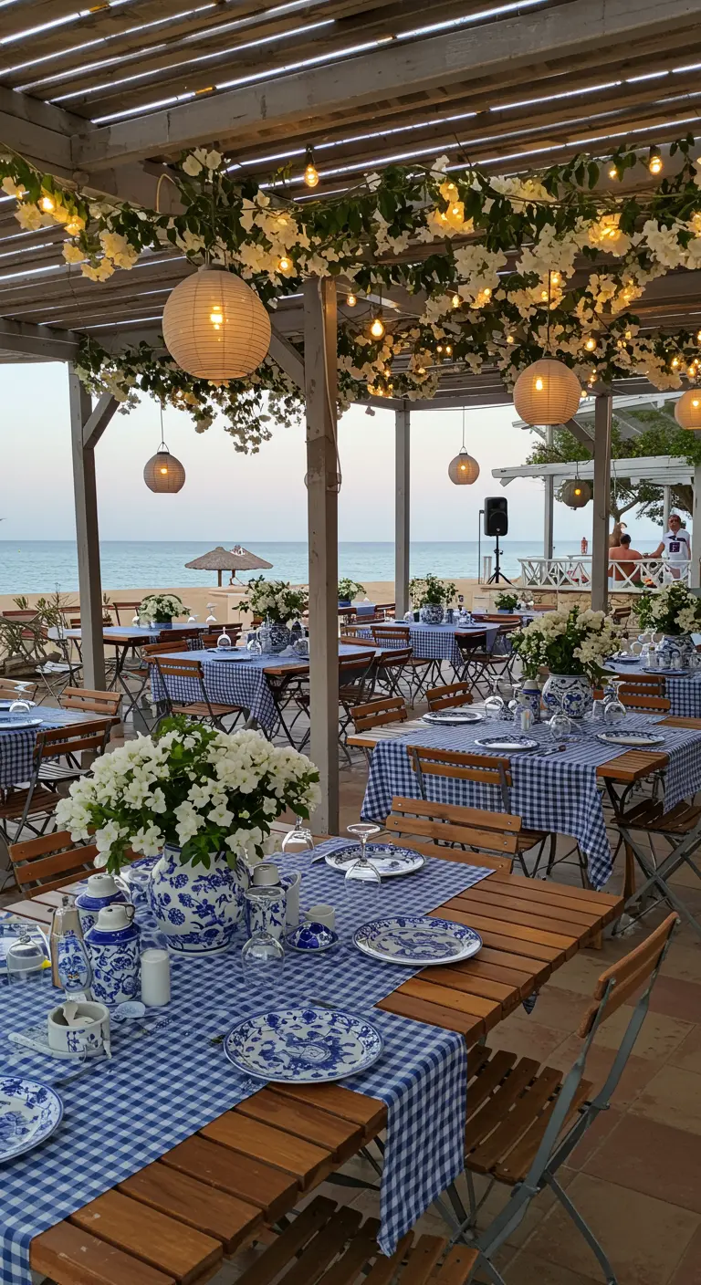 Restaurant de plage avec tables sous pergola fleurie, nappes vichy bleues, et lampions suspendus.