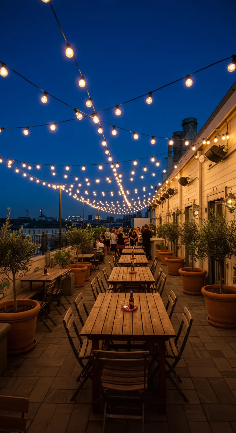 Terrasse urbaine avec de nombreuses guirlandes lumineuses, tables en bois, et pots d'oliviers, ambiance guinguette.