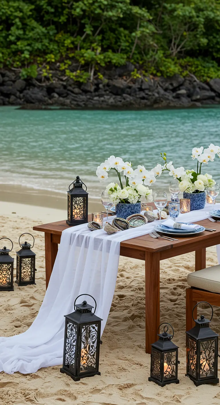 Table de dîner sur la plage avec lanternes noires, fleurs blanches dans des vases bleus et coquillages.